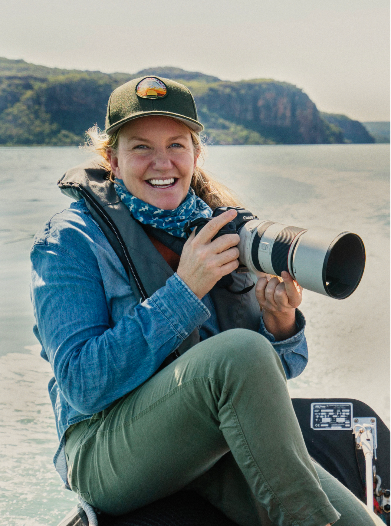 Photo of Elise Hassey. She is smiling and holding a camera with a large lens. In the background is ocean and cliffs.