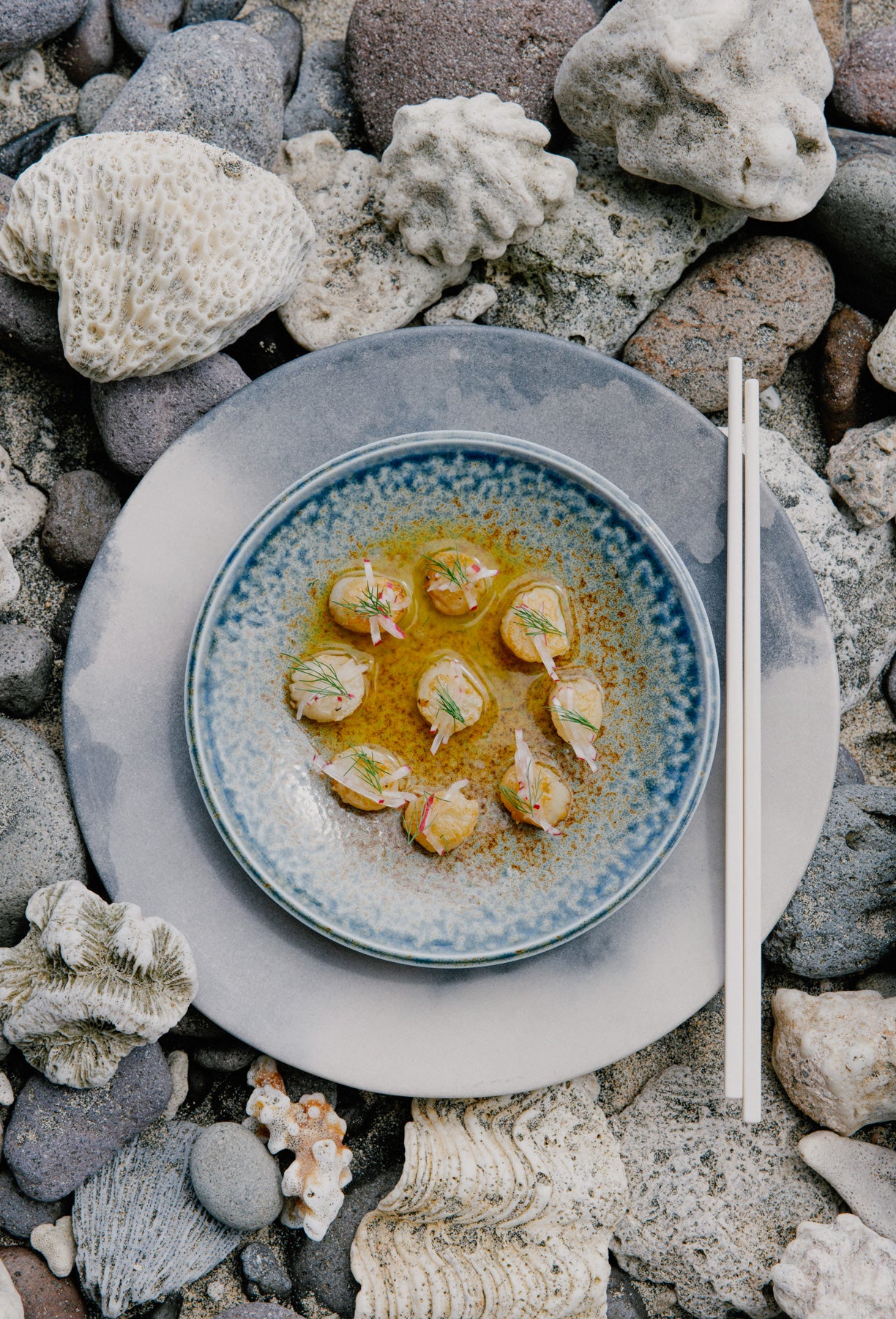 A gourmet seafood dish in a blue ceramic bowl surrounded by coral and stone on a beach