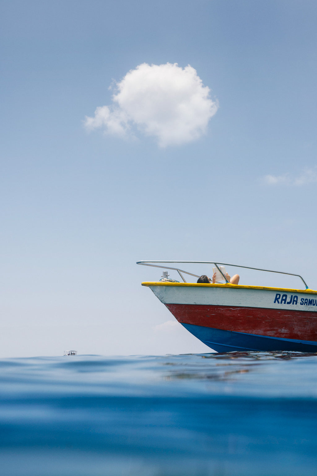 Colourful fishing boat floating on calm sea with woman sunbathing and a single cloud above