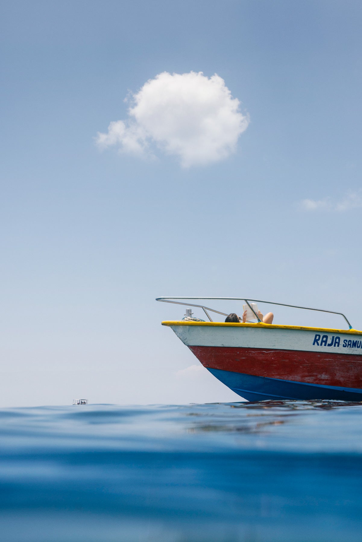 Colourful fishing boat floating on calm sea with woman sunbathing and a single cloud above