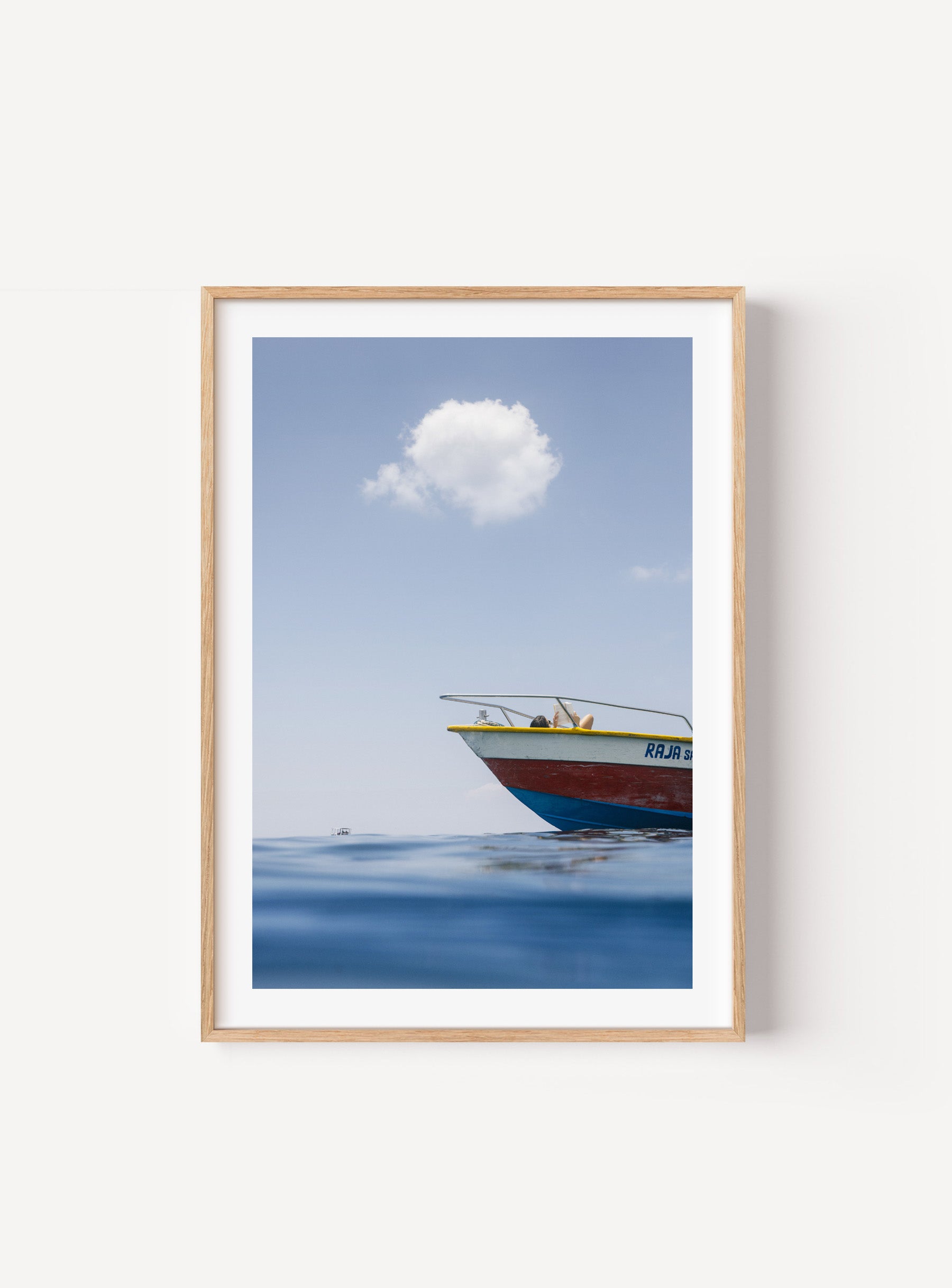Colourful fishing boat floating on calm sea with woman sunbathing and a single cloud above, mocked up in a frame on a wall