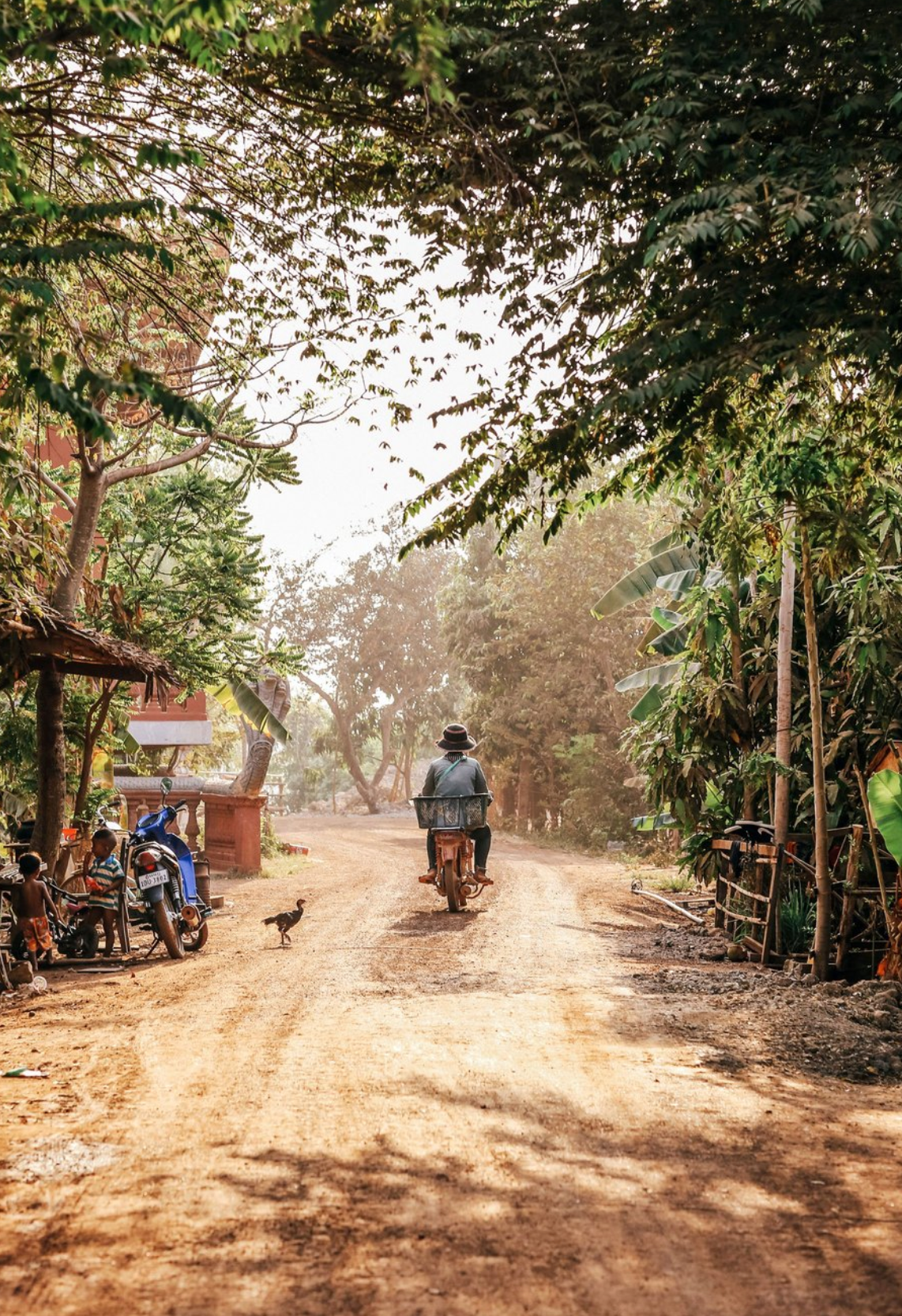 Man riding motorbike down a dusty village road in Cambodia with children and tropical trees