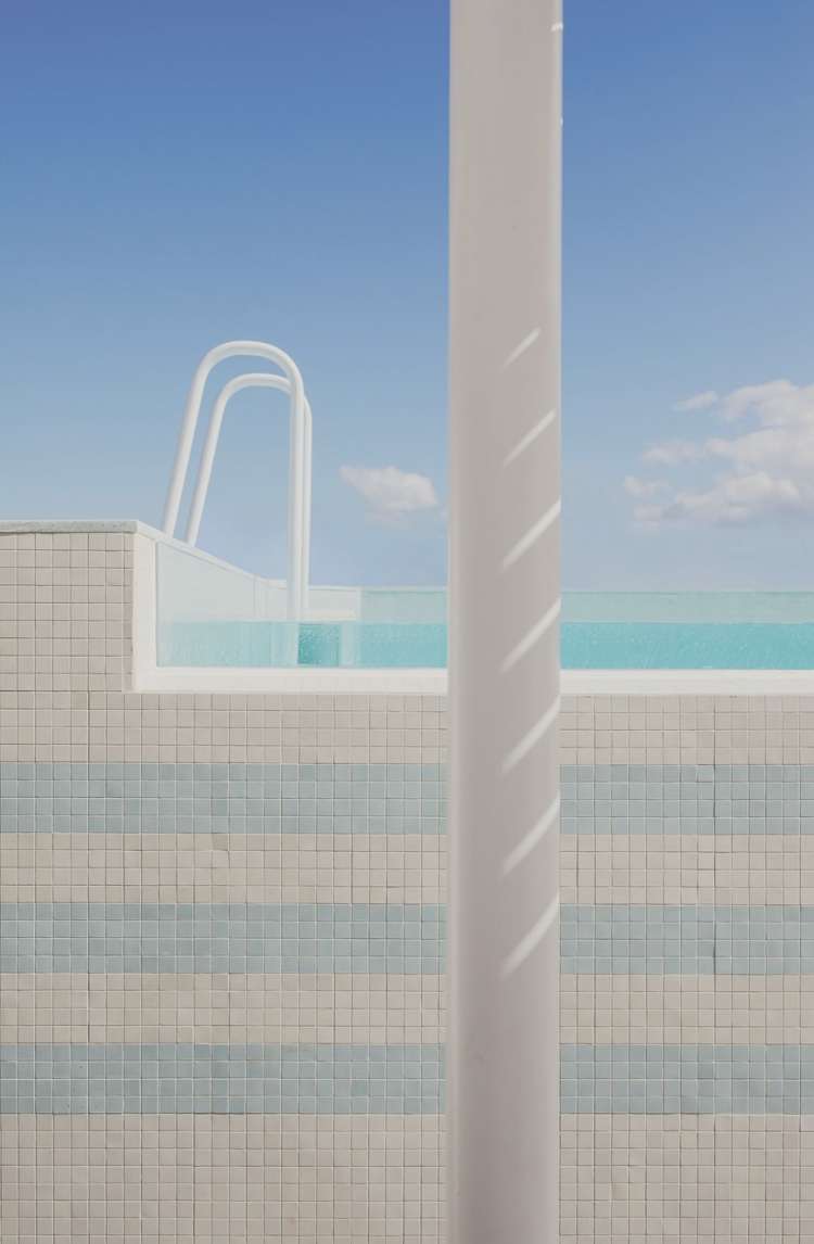 Tiled rooftop pool with white ladder and striped mosaic wall, set against a clear blue sky with soft clouds